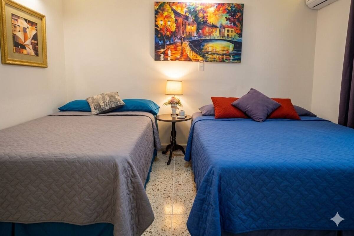 A hotel room with two beds, a nightstand with a lamp, a painting on the wall, and a small table with a vase.