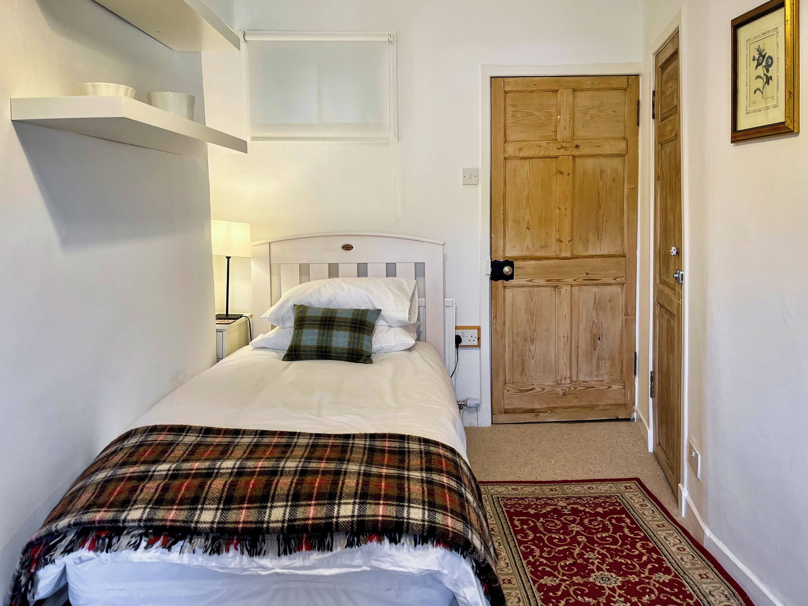 A neatly made bed with a plaid blanket, a bedside lamp, and a wooden door in a simple room with white walls.