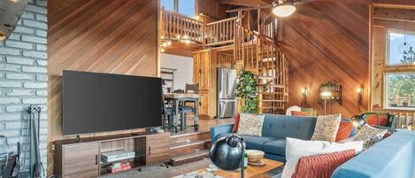 A spacious loft-style living area with a large flat-screen TV, a blue sofa, wooden flooring, and a staircase leading to an upper level.
