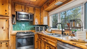 A kitchen with wooden cabinets, stainless steel appliances, a green tiled backsplash, and a window with a view of trees.