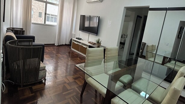 A modern living room with a glass dining table, comfortable seating, a flat-screen TV, and a large window with curtains.