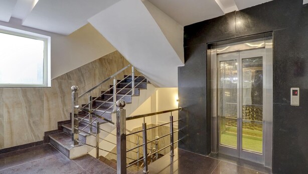 A modern interior with a marble staircase, a glass elevator, and a view of a green outdoor area. - Treebo Trend Hotel NV (Narendranagar)