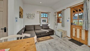 A cozy living room with a sofa, a wooden table, and a door leading outside. - Lodge in Suffolk Countryside With BBQ (Bury St Edmunds)