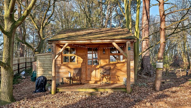 A wooden cabin with a porch, two chairs, and a small table. - Lodge in Suffolk Countryside With BBQ (Bury St Edmunds)