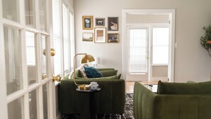 Quarto com dois sofás verdes, uma pequena mesinha lateral preta e um candeeiro dourado. Há quadros nas paredes e uma porta com persianas brancas.