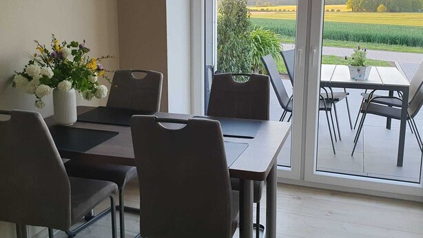 Een eethoek met een tafel en stoelen, een bloemenvaas en uitzicht op een veld door grote glazen deuren.