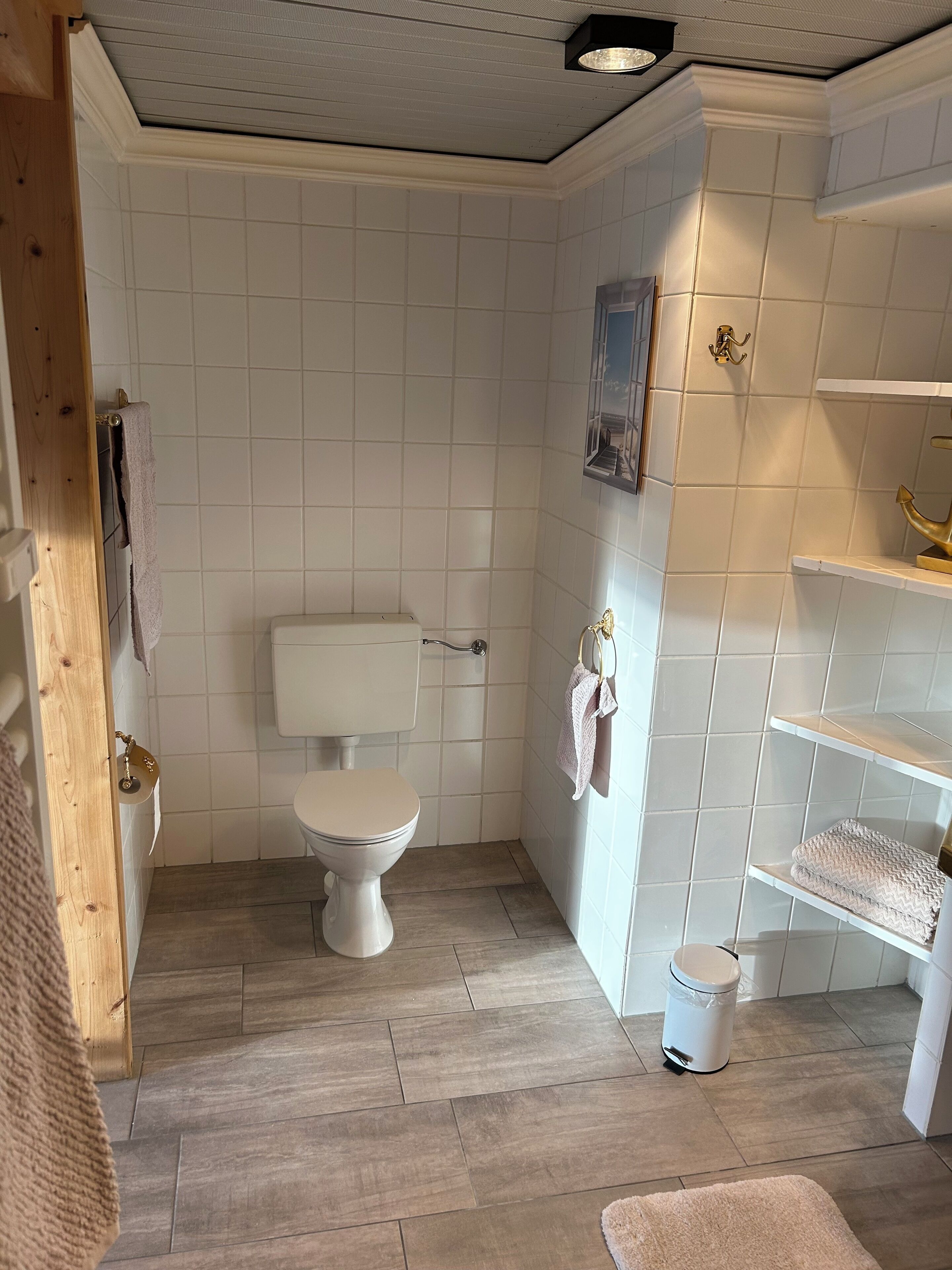 A bathroom with white tiled walls, a white toilet, a wooden floor, and a small trash bin.