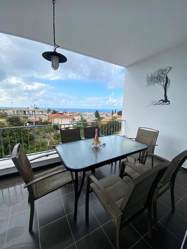 Un balcón con una mesa y sillas de vidrio, una lámpara colgante y vistas a la ciudad y al mar.