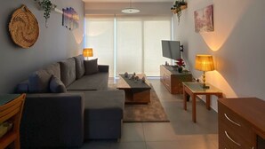 Un salon moderne avec un canapé gris, une table basse et un téléviseur. Des plantes ornent les murs et une lampe est posée sur une table d’appoint.