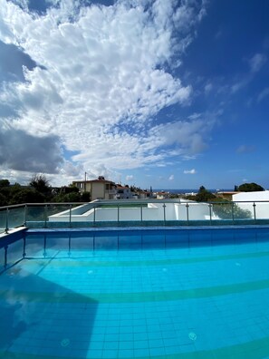A swimming pool with clear blue water, surrounded by a glass railing. In the background, there are buildings and a view of the sea under a partly cloudy sky.