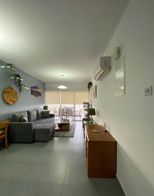 Una sala moderna con un sofá seccional gris, una mesa de centro de madera y una mesita auxiliar de madera. Se ve un balcón con puerta de vidrio al fondo.