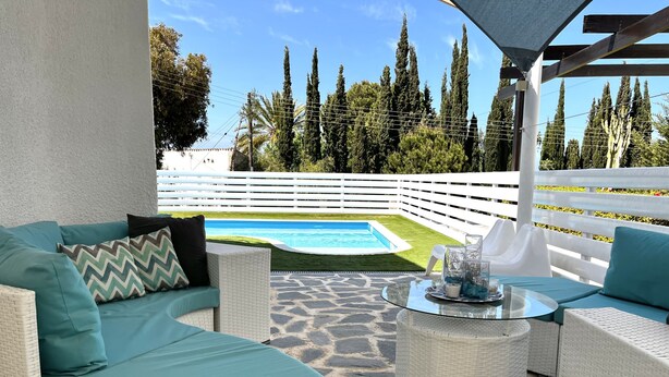 Un'area patio con mobili in vimini, un tavolo in vetro e vista su una piscina.