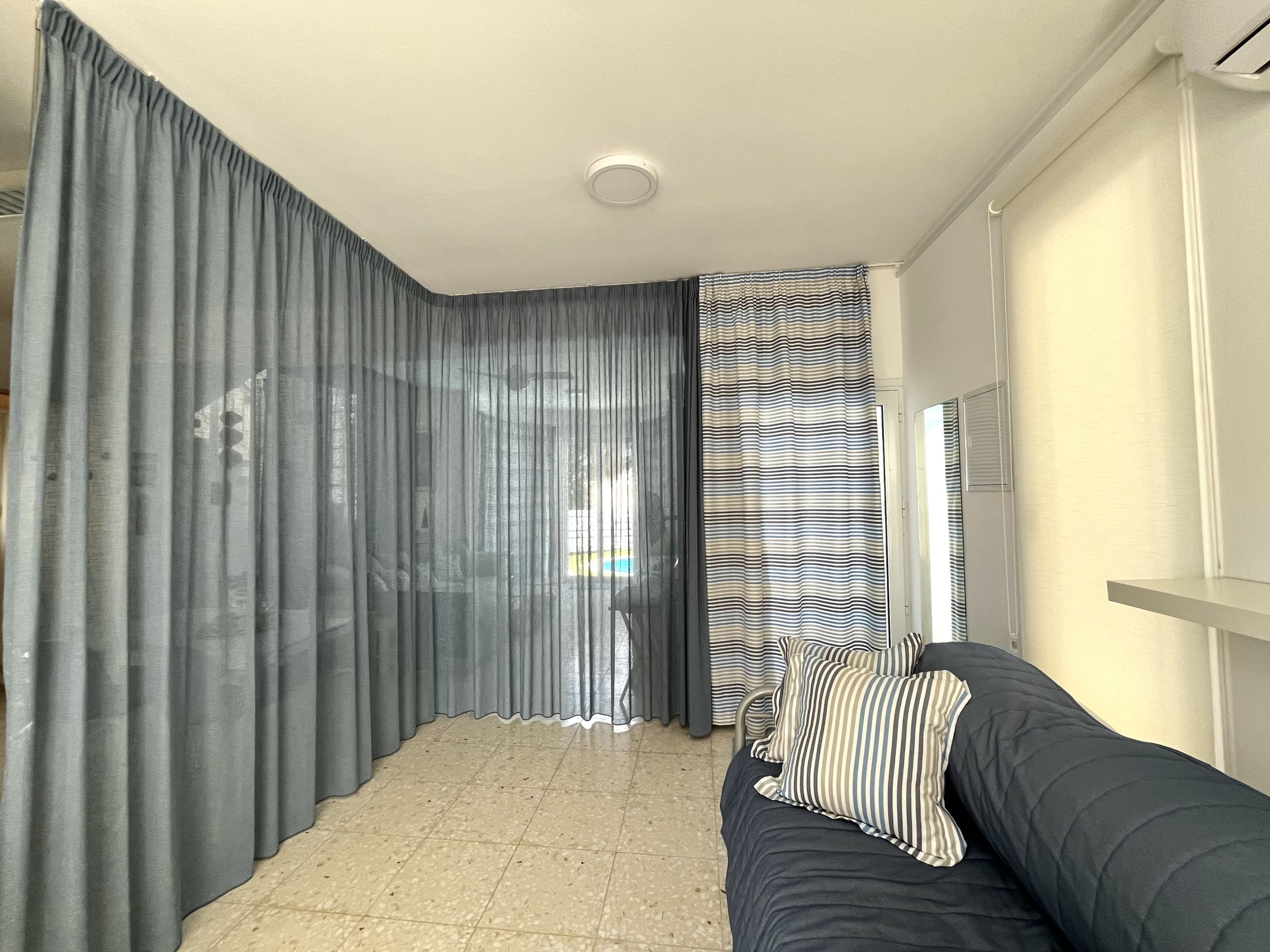 Una habitación con un sofá de color oscuro, una cortina a rayas y suelo de baldosas.