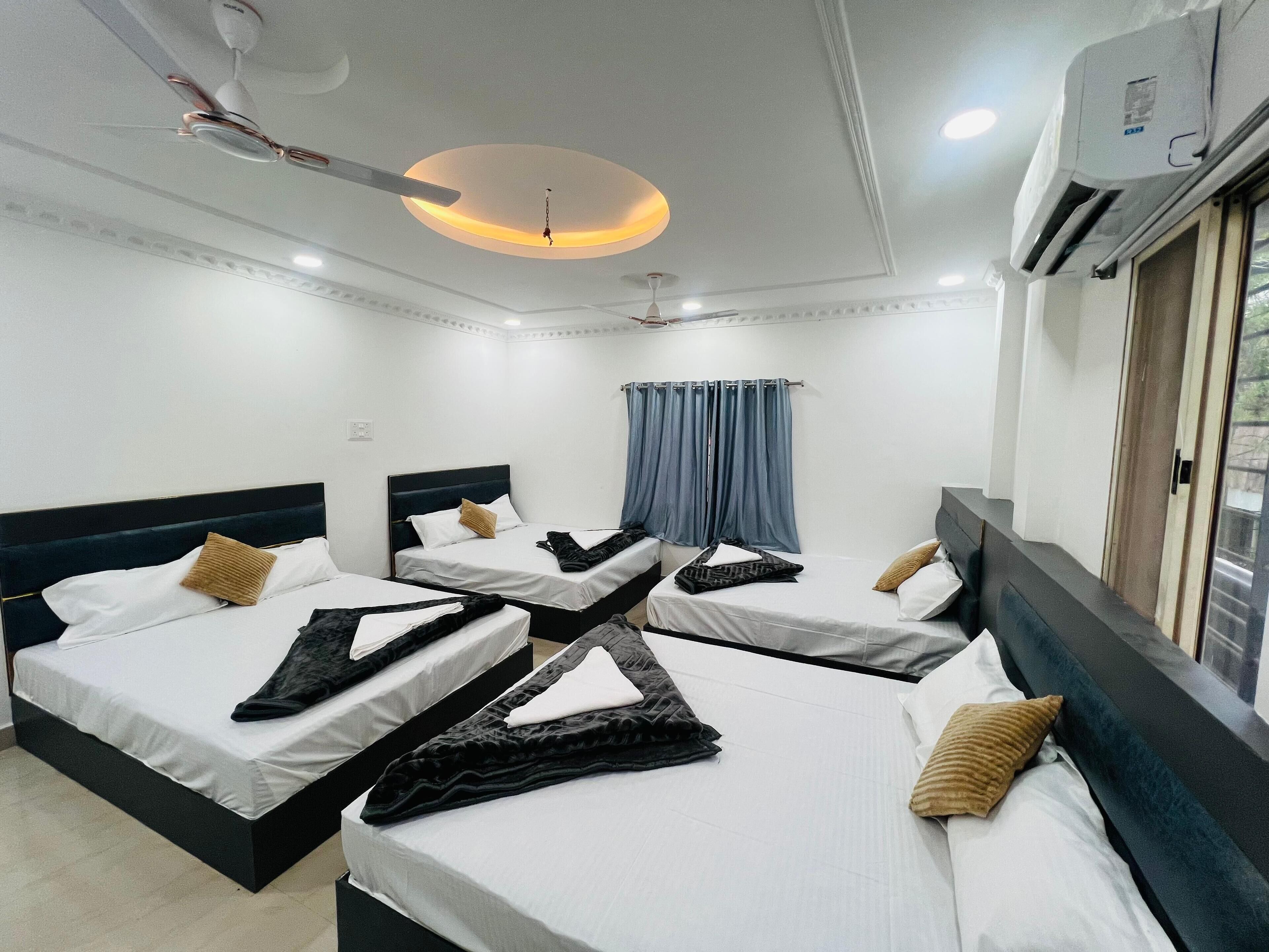 A hotel room with four beds arranged in a square layout, each with white linens and dark headboards. There is a ceiling fan, an air conditioning unit, and a window with curtains.