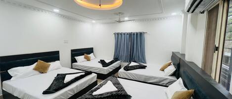 Et hotelværelse med fire senge arrangeret i en firkant, hver med hvide lagner og mørke sengegærder. Der er en ventilator, en aircondition og et vindue med gardiner.