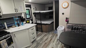 A compact kitchenette with a stove, oven, and sink, a bunk bed, a small table with a chair, and a clock on the wall.