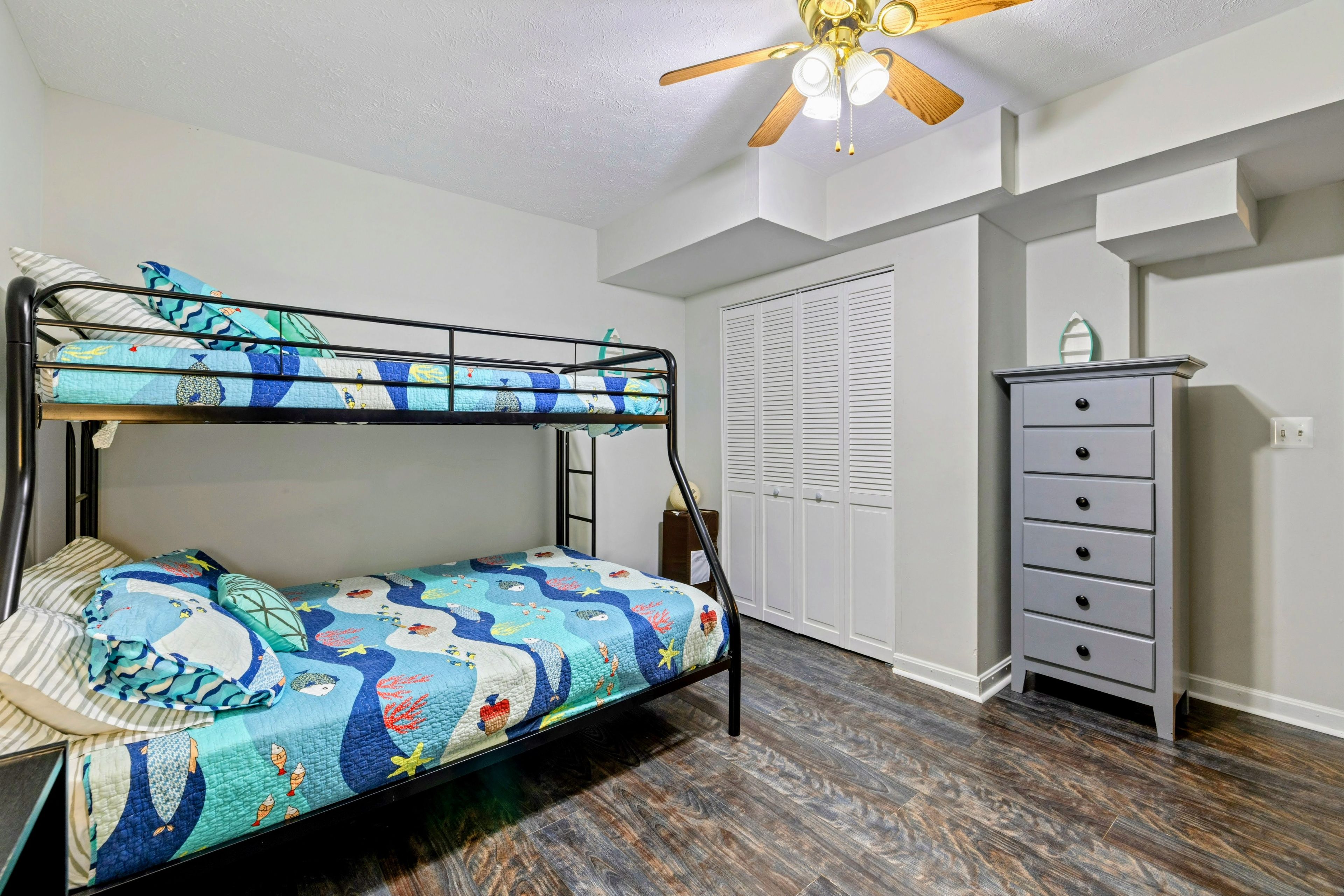 Un lit superposé avec une courtepointe aux couleurs vives, une commode et un ventilateur de plafond dans une pièce au plancher en bois et aux murs blancs.