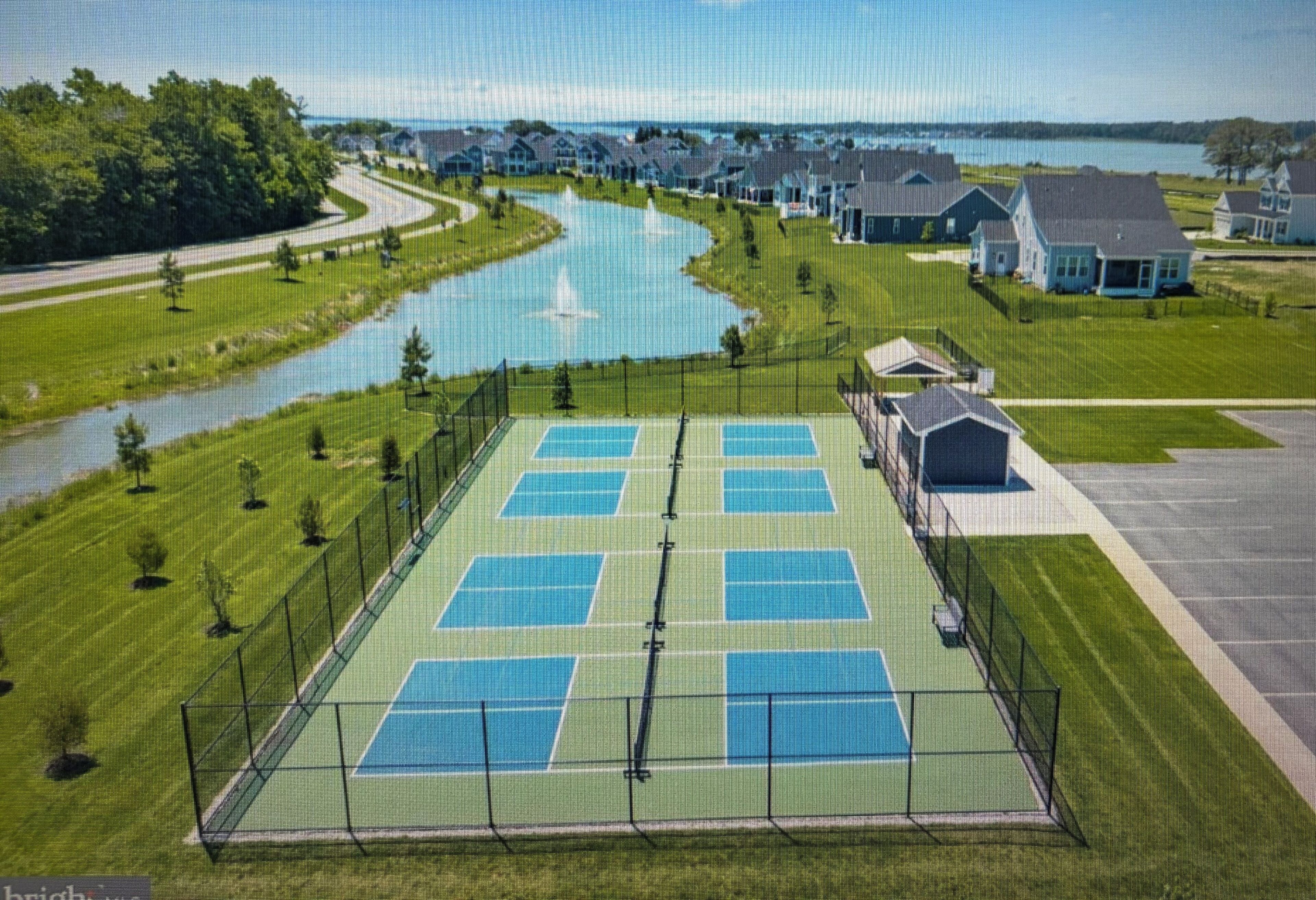 Ein Tennisplatz, umzäunt, mit Häusern und einem Gewässer im Hintergrund.