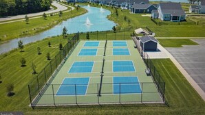 A tennis court surrounded by a fence, with houses and a body of water in the background.