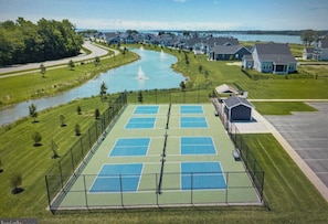A tennis court surrounded by a fence, with houses and a body of water in the background.