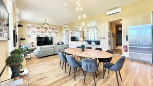 A spacious living room with a large TV, a dining area with a wooden table and chairs, and a kitchen area with a refrigerator.
