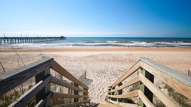Ein Holzsteg führt zu einem Strand, im Hintergrund ist ein Steg zu sehen.