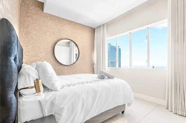 A modern hotel room with a large bed, a round mirror, a view of the city, and a large window with curtains.