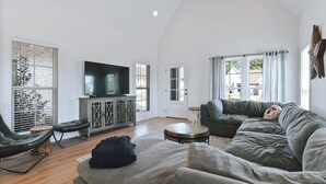 A spacious living room with a grey sofa, a black leather chair, a television on a cabinet, and a large window with curtains. - Charming 2-bedroom townhome with WiFi and AC in Auburn (Auburn)