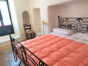 Un lit avec un cadre en métal et une couverture rose, une chaise, une lampe et une fenêtre donnant sur un paysage verdoyant.