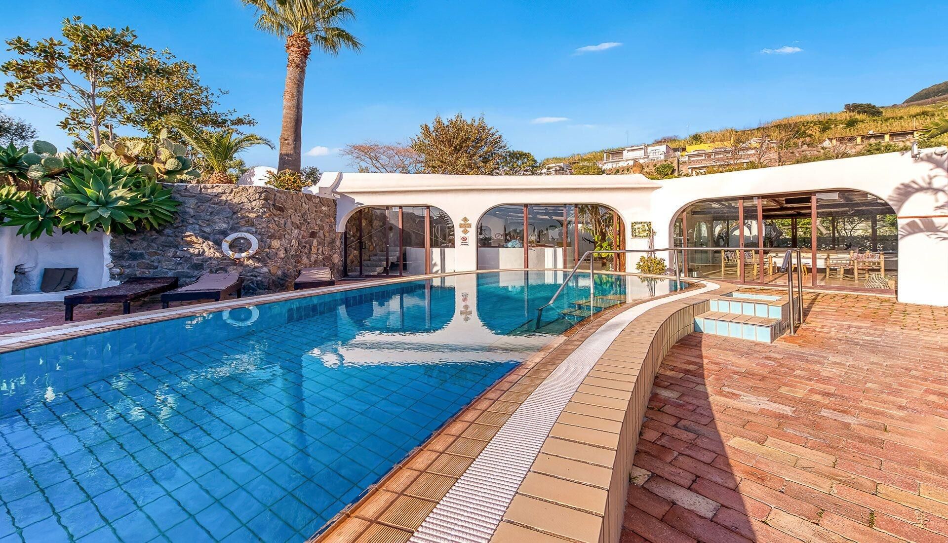A swimming pool with a tiled deck, surrounded by stone walls and palm trees.