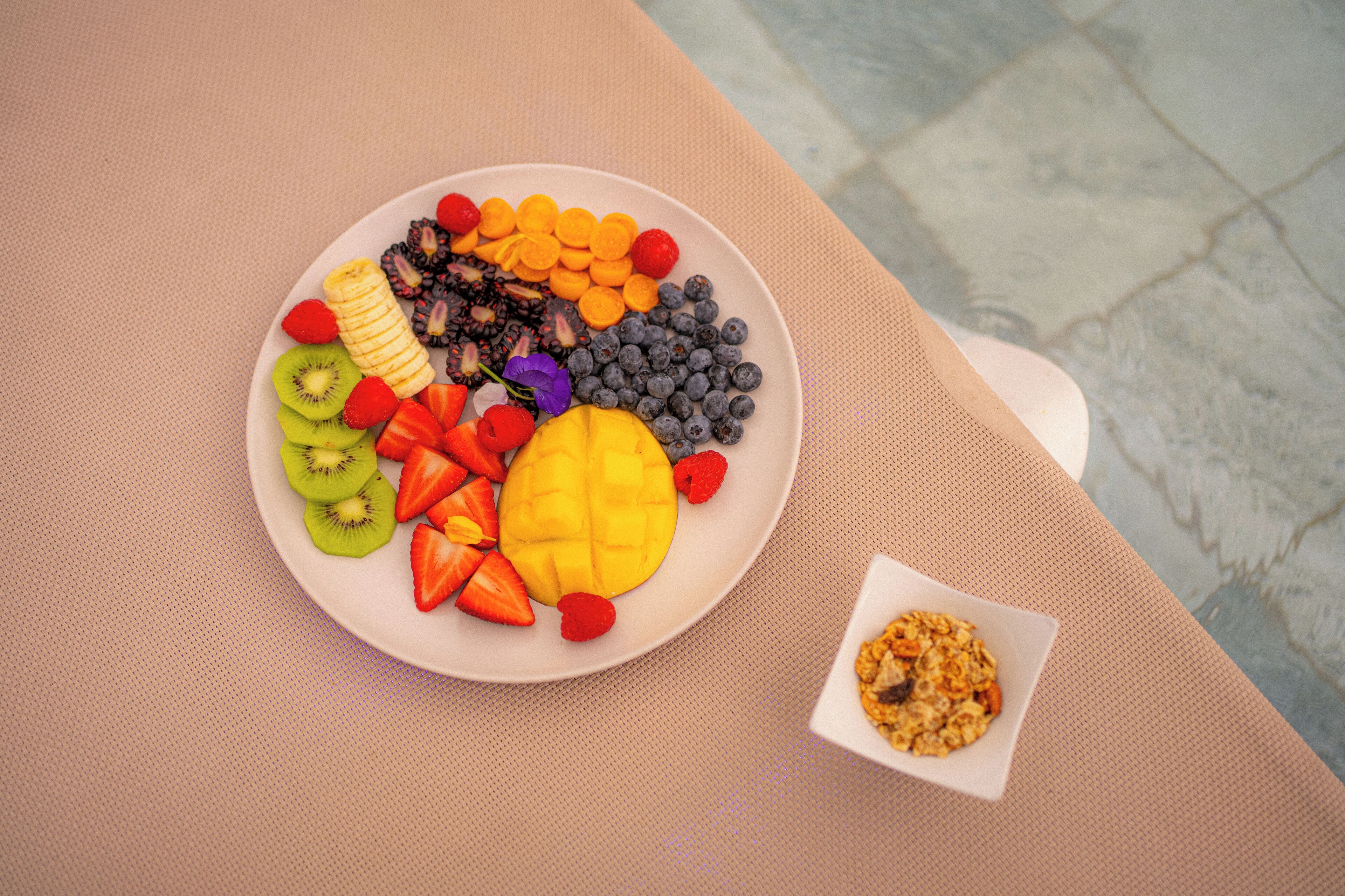 Un plato de frutas variadas y un cuenco pequeño de granola sobre una mesa con textura.