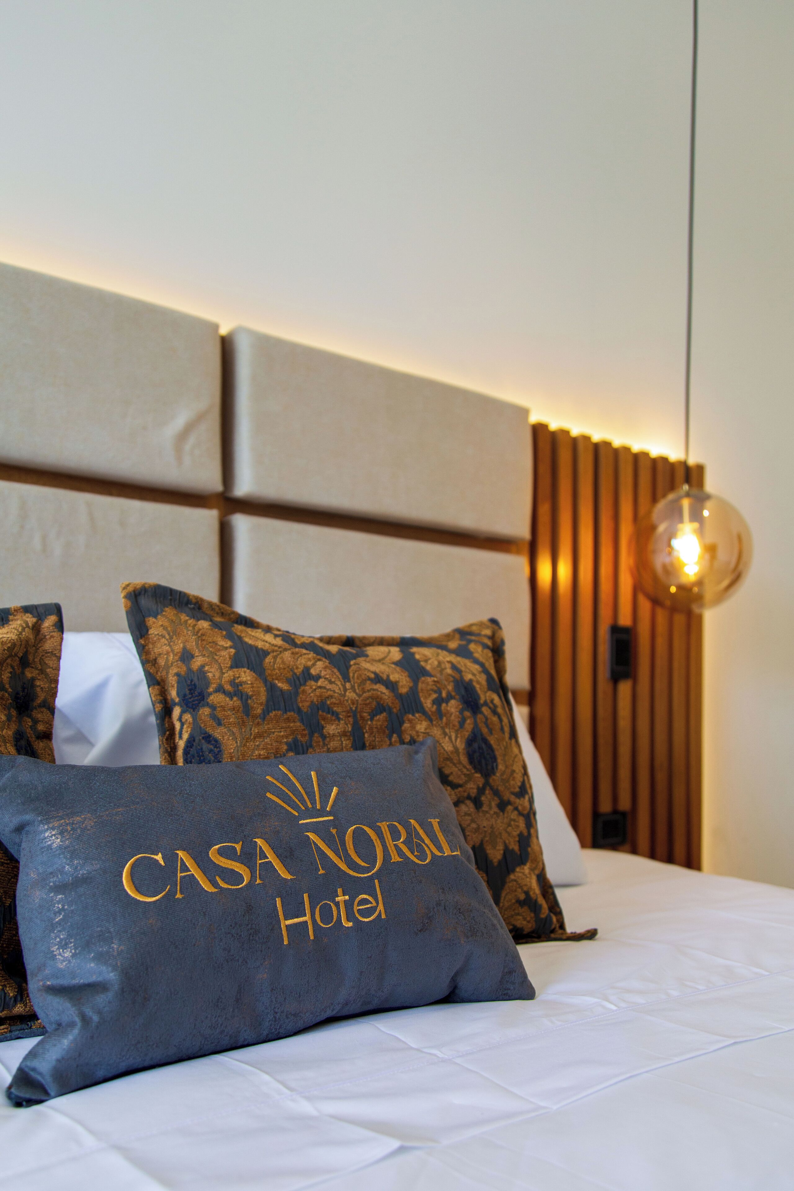 A neatly made bed with a pillow labeled "CASA NORAL Hotel" and a wooden headboard with vertical slats.