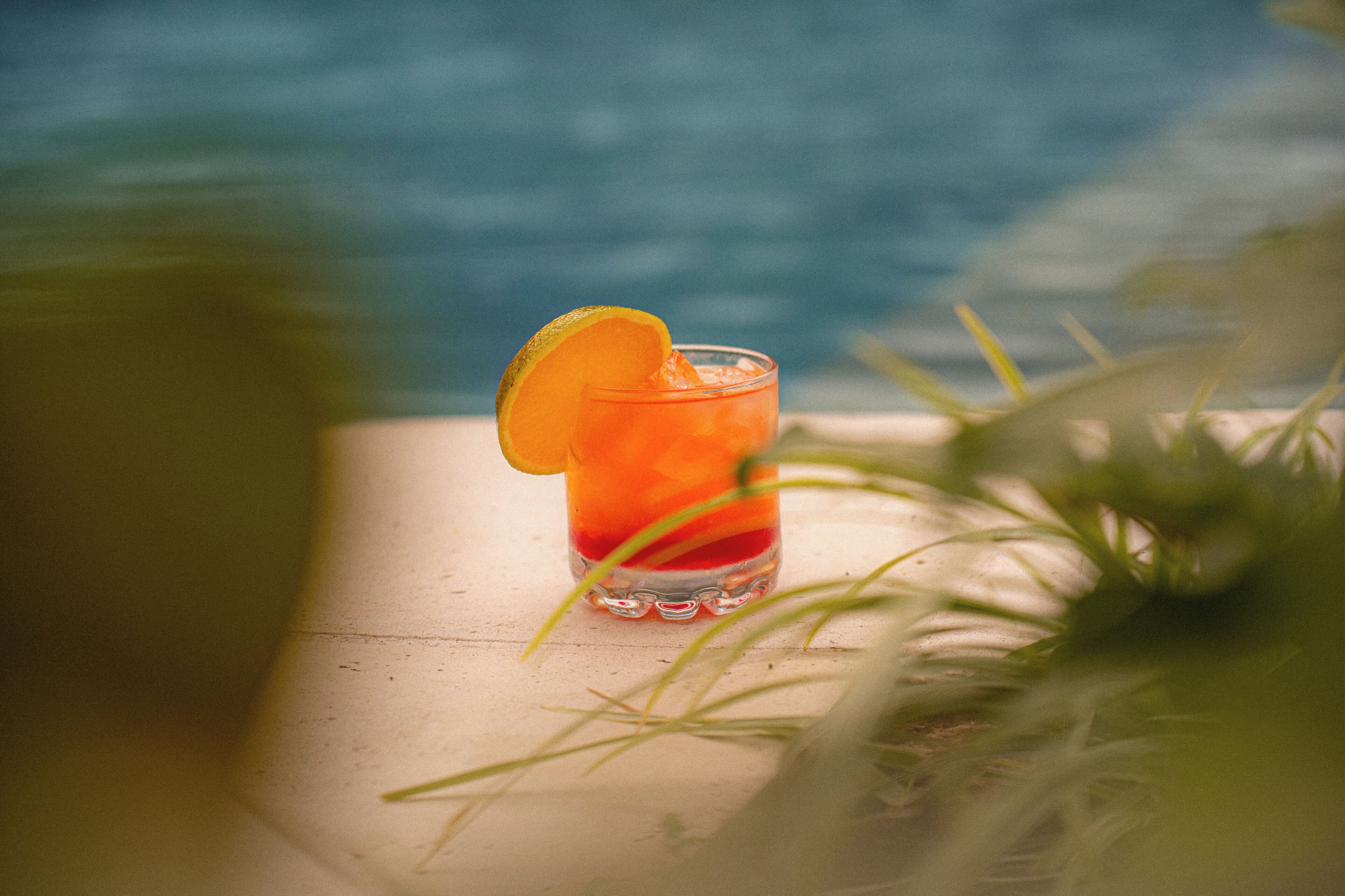 Un verre d’une boisson rouge, agrémenté d’une tranche d’orange, posé sur une surface en bois près d’une piscine.
