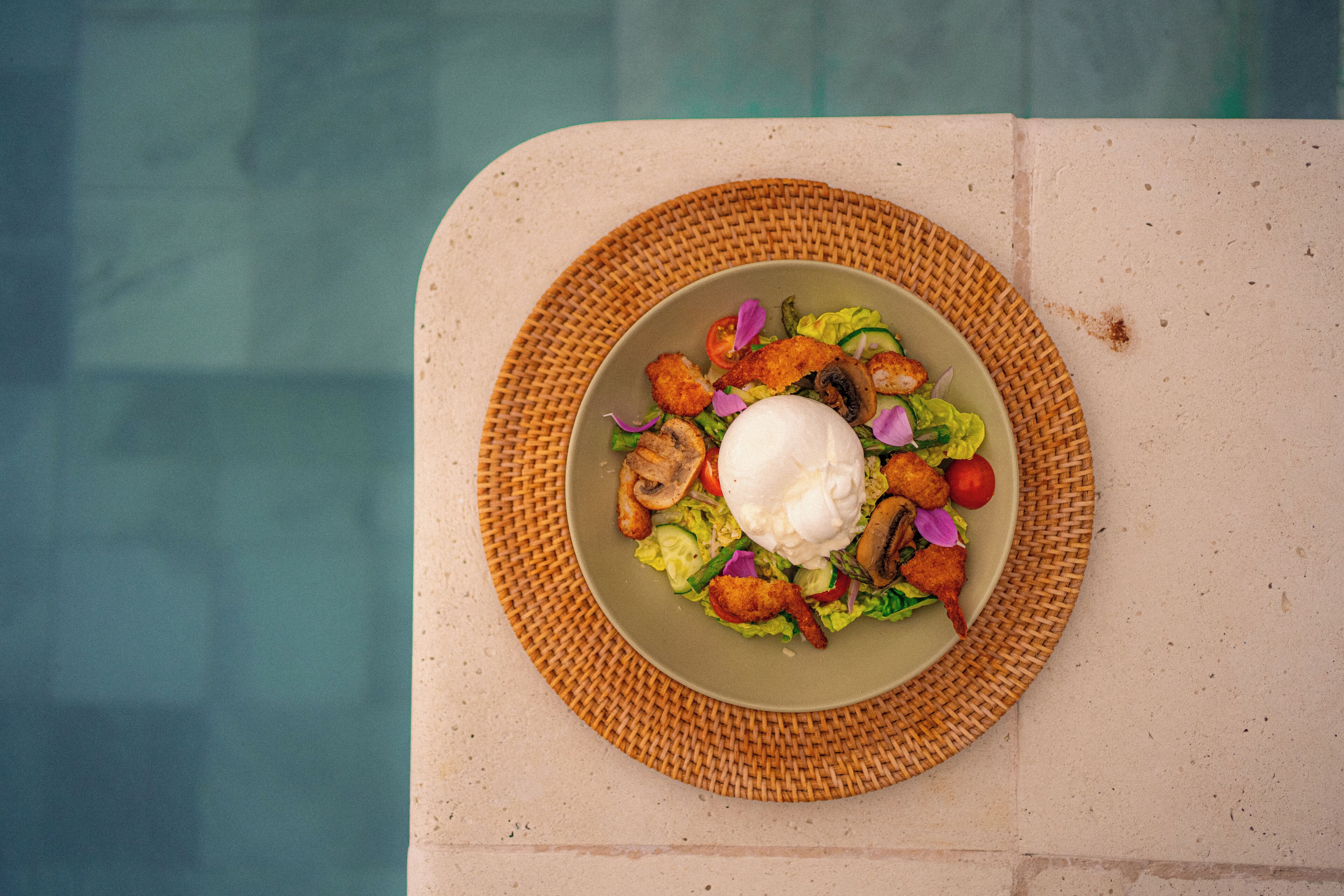 Un plato de comida con un huevo pochado, champiñones y verduras sobre una estera tejida junto a una piscina.