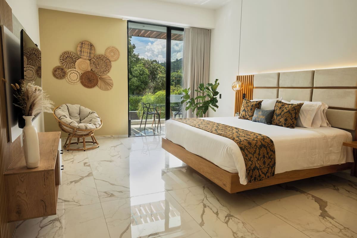 A modern bedroom with a large bed, a wicker chair, a TV, and a balcony with a view of greenery.