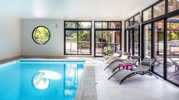An indoor swimming pool with clear glass walls, lounge chairs, and a small table. - Ra Sid Spa Loire Sa Vre (Reze)
