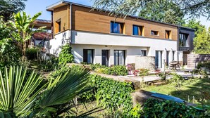 Una moderna casa a due piani con facciata in legno, ampie finestre e un giardino curato con diverse piante e un piccolo patio.