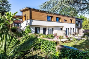 A modern two-story house with a wooden facade, large windows, and a well-maintained garden with various plants and a small patio area. - Ra Sid Spa Loire Sa Vre (Reze)