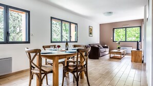 A spacious living area with a wooden dining table and chairs, a sofa, and a TV stand. Large windows provide natural light and views of greenery.