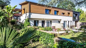 A modern two-story house with a wooden facade, large windows, and a well-maintained garden with various plants and a small patio area. - Ra Sid Spa Loire Sa Vre (Reze)