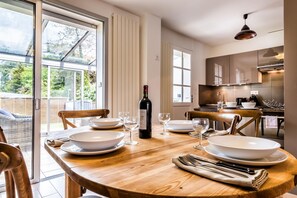 A dining area with a wooden table set for four, a bottle of wine, and a view of the garden through large windows. - Ra Sid Spa Loire Sa Vre (Reze)