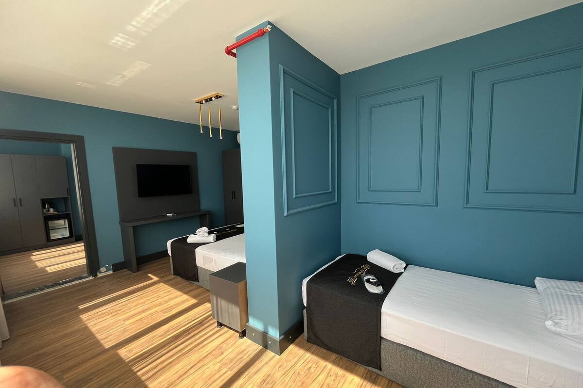 A modern hotel room with a large bed, a smaller bed, a desk with a chair, a TV mounted on the wall, and a wardrobe.