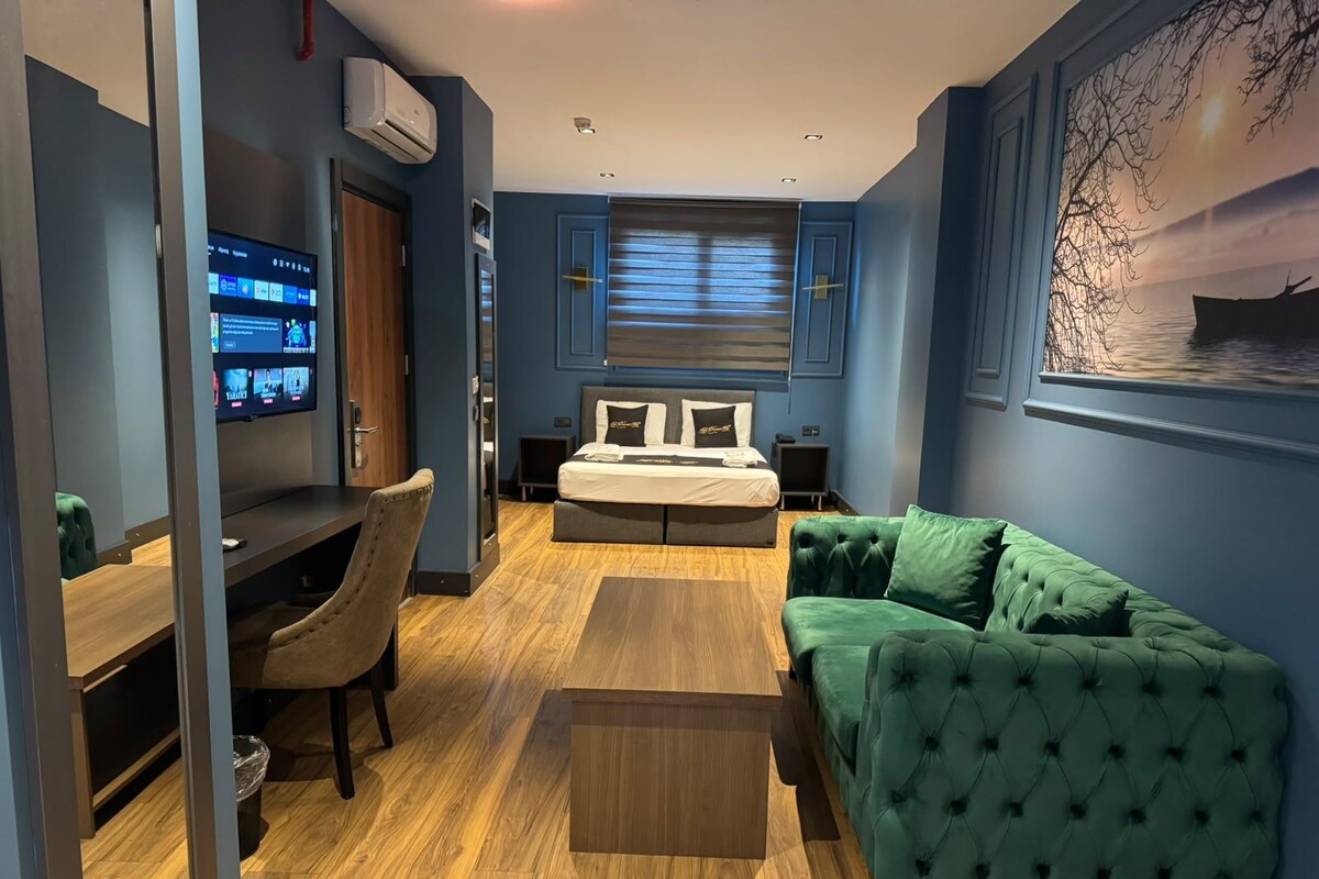 A modern hotel room with a bed, a green sofa, a desk, and a TV mounted on the wall.