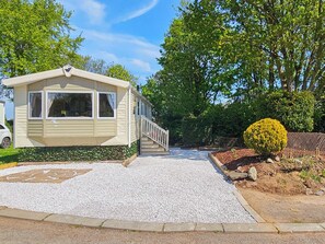 A mobile home with a gravel driveway, a small porch, and a well-maintained garden with a hedge and a bush.