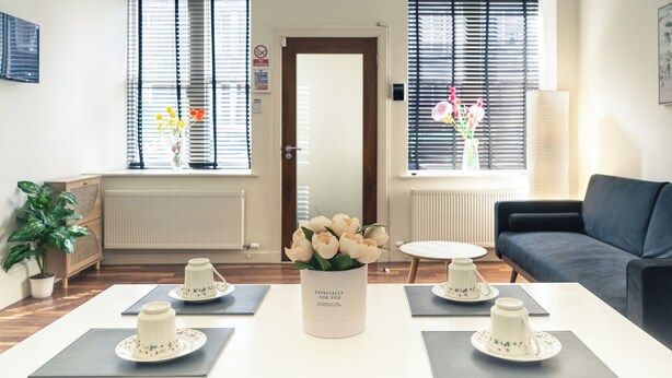 A modern living room with a sofa, a coffee table, and a vase of flowers. There are two windows with blinds, a door, and a radiator. - Entire Family Apartment- Edinburgh (Edinburgh)