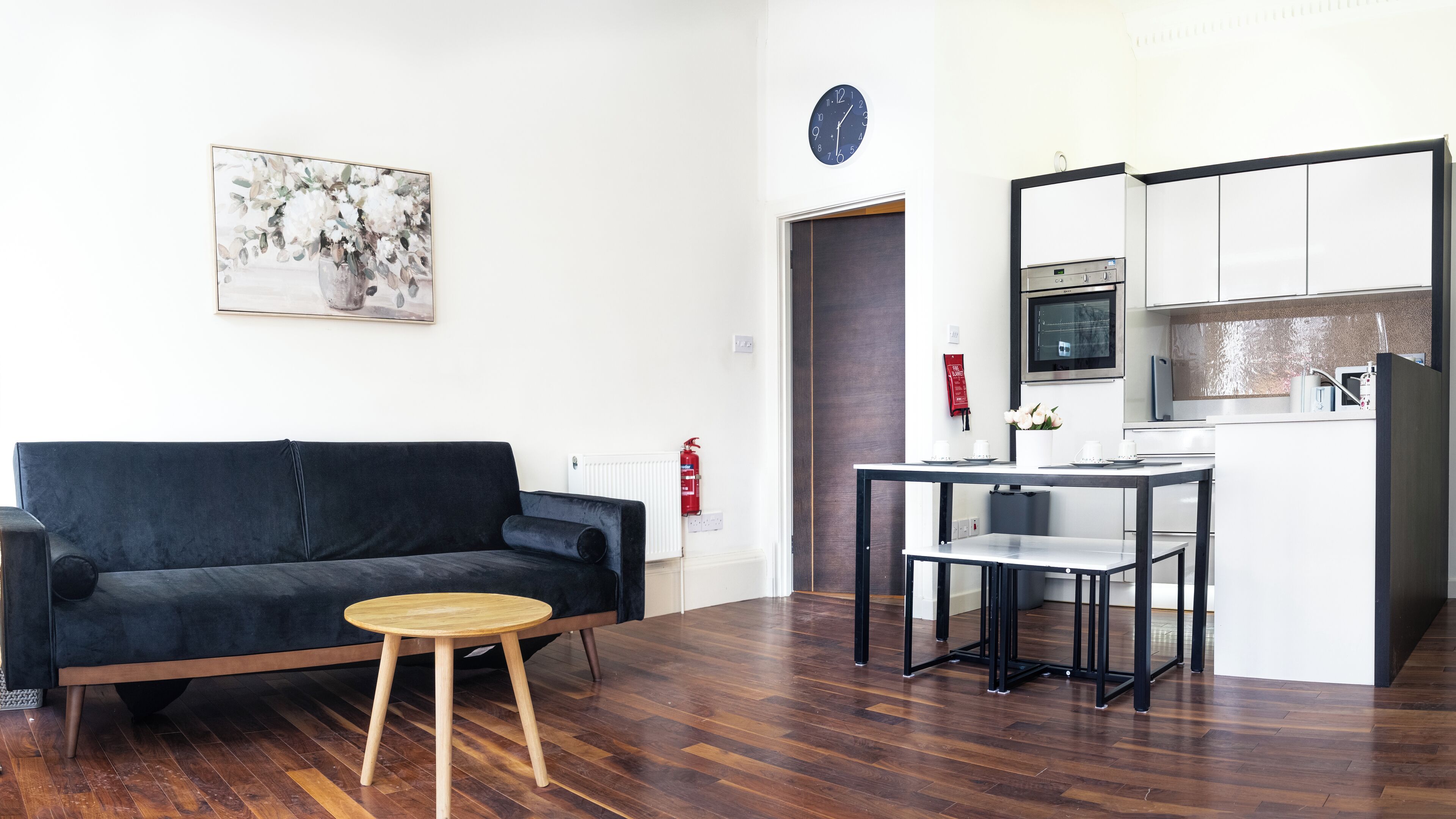 A modern living room with a dark sofa, a small round wooden table, and a kitchen area with a microwave, oven, and sink.