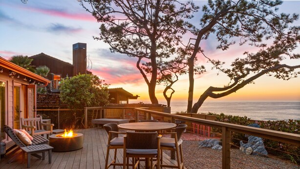 A wooden deck with a round table and chairs, a fire pit, and a view of the ocean at sunset.