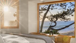 A bedroom with a large window overlooking a beach, a bed, a chair, and a table with a pitcher.