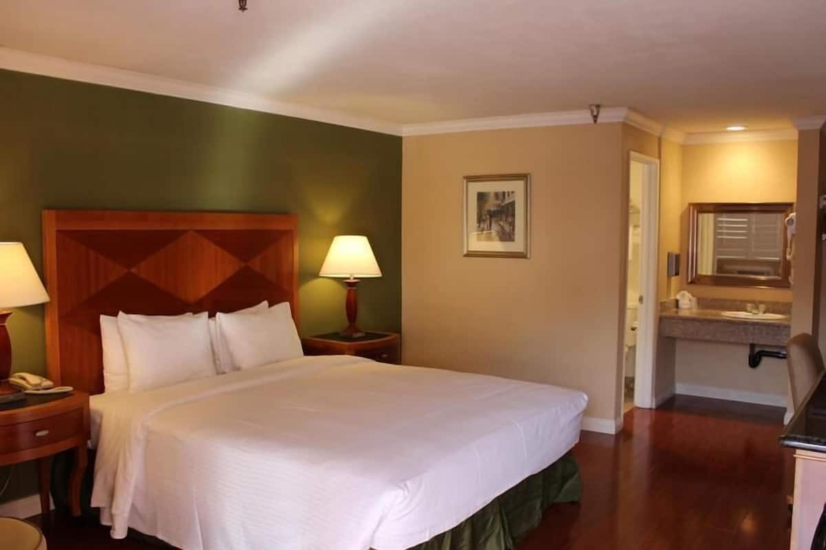 A hotel room with a large bed, two bedside tables with lamps, a headboard, a framed picture on the wall, and a bathroom with a sink and mirror.