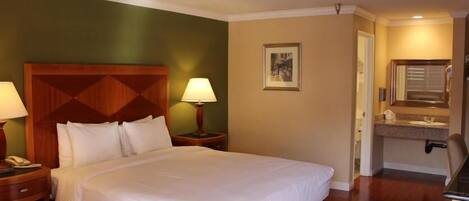 Habitación de hotel con una cama grande, dos mesitas de noche con lámparas, un cabecero, un cuadro enmarcado en la pared y un baño con lavabo y espejo.